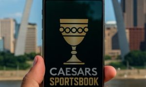 Realistic photo of a hand holding a smartphone showing the Caesars goblet logo on the screen, with the St. Louis Gateway Arch in the background.