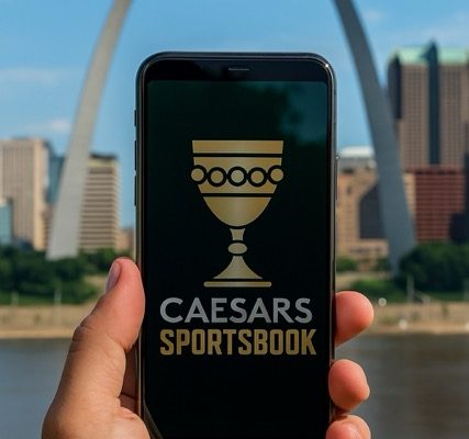 Realistic photo of a hand holding a smartphone showing the Caesars goblet logo on the screen, with the St. Louis Gateway Arch in the background.
