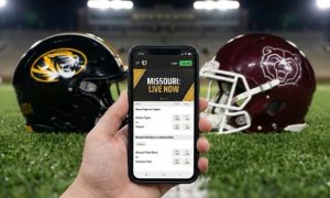 A first-person view of a hand holding a smartphone displaying a Missouri sports betting app with live odds, set against a background of Mizzou and Missouri State football helmets on a stadium field.