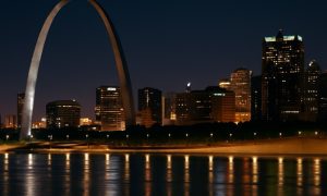 St. Louis skyline at night with the Gateway Arch illuminated above the Mississippi River, reflecting warm city lights on the water.