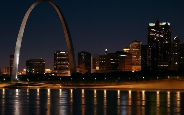 St. Louis skyline at night with the Gateway Arch illuminated above the Mississippi River, reflecting warm city lights on the water.
