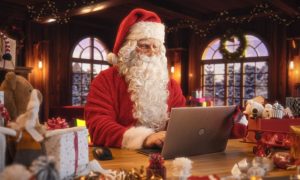 Santa Sits and Plays On Laptop Inside His House Surrounded By Christmas Gifts