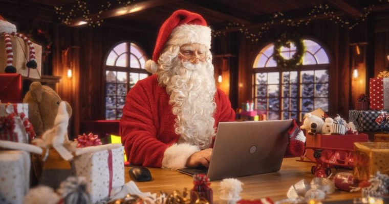 Santa Sits and Plays On Laptop Inside His House Surrounded By Christmas Gifts
