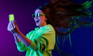Young Happy Woman Looks At Cell Phone With Flying Hair and Neon Sunglasses