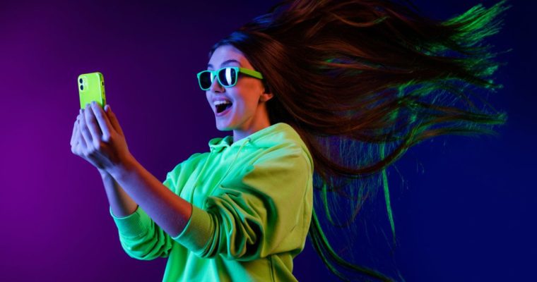 Young Happy Woman Looks At Cell Phone With Flying Hair and Neon Sunglasses