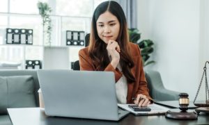 Woman Sits Thinking in Front of Computer with Gavel, Scale and Calculator on Desk