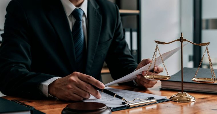 Man Holding Pen Studies Papers Next to Scale and Judge's Gavel on Desk