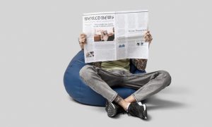 Man Sits with Open Legs on Bean Bag Reading Newspaper on Gray Background