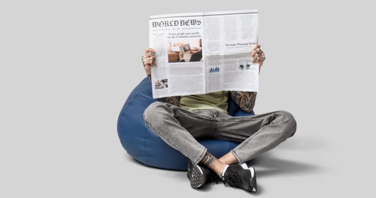 Man Sits with Open Legs on Bean Bag Reading Newspaper on Gray Background