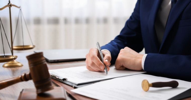 Man In Suit Writes On Documents In Office, On Desk Sits Scale and Judges Gavel