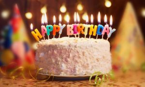 White Cake with Lit Candles Spelling Out Happy Birthday On Table with Party Hats Background
