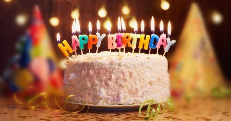 White Cake with Lit Candles Spelling Out Happy Birthday On Table with Party Hats Background