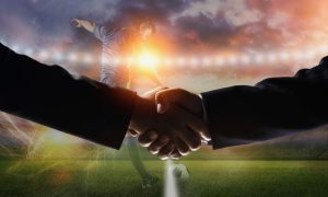 Soccer Player Kicks Ball on Field with Two Men Shaking Hands in Foreground