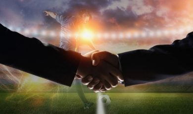 Soccer Player Kicks Ball on Field with Two Men Shaking Hands in Foreground