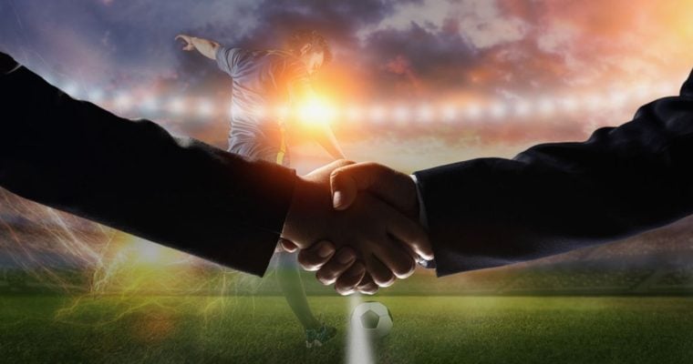 Soccer Player Kicks Ball on Field with Two Men Shaking Hands in Foreground
