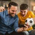 Young Male and Older Male Look Excitedly at Cell Phone While Holding Soccer Ball in Living Room