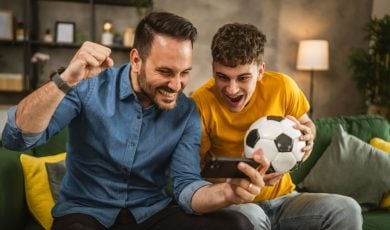 Young Male and Older Male Look Excitedly at Cell Phone While Holding Soccer Ball in Living Room