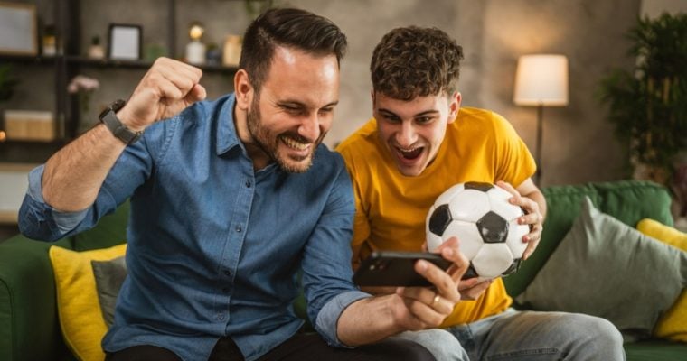 Young Male and Older Male Look Excitedly at Cell Phone While Holding Soccer Ball in Living Room