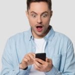 Young Man Looks At His Cell Phone With Surprised Expressioin on Isolated Background