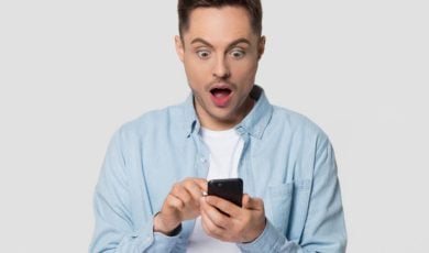 Young Man Looks At His Cell Phone With Surprised Expressioin on Isolated Background