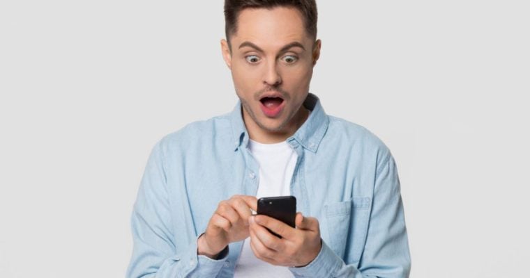 Young Man Looks At His Cell Phone With Surprised Expressioin on Isolated Background