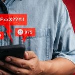 Man in Denim Shirt Holds Cell Phone Displaying Negative Comments in Red
