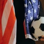 Soccer Fan Holds Ball in One Hand Wearing Black T-Shirt and American Flag Around Shoulders