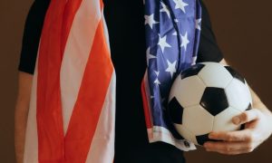 Soccer Fan Holds Ball in One Hand Wearing Black T-Shirt and American Flag Around Shoulders