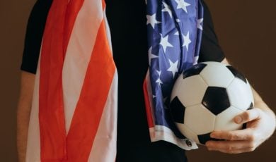 Soccer Fan Holds Ball in One Hand Wearing Black T-Shirt and American Flag Around Shoulders