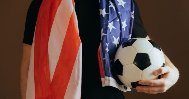 Soccer Fan Holds Ball in One Hand Wearing Black T-Shirt and American Flag Around Shoulders