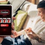 Happy Man with Tattoos Sits On Couch and Cell Phone Displaying Online Casino in Foreground