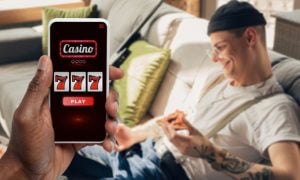 Happy Man with Tattoos Sits On Couch and Cell Phone Displaying Online Casino in Foreground