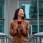 Young Asian Woman Stands In Front of Building Holding Cell Phone Smiling