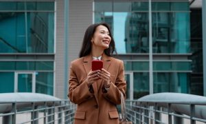 Young Asian Woman Stands In Front of Building Holding Cell Phone Smiling