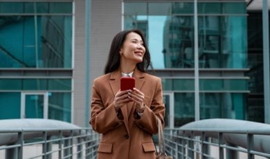 Young Asian Woman Stands In Front of Building Holding Cell Phone Smiling