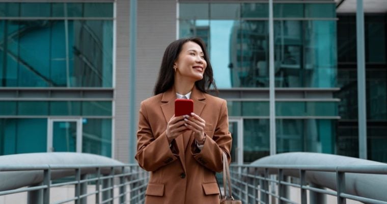 Young Asian Woman Stands In Front of Building Holding Cell Phone Smiling