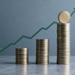 Three stacks of gold coins of increasing height are arranged in front of an upward-trending green line graph against a grey background.
