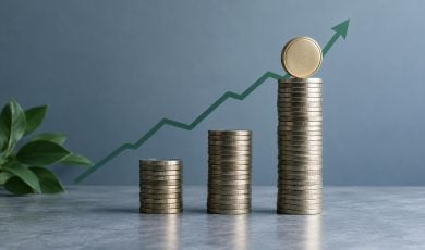 Three stacks of gold coins of increasing height are arranged in front of an upward-trending green line graph against a grey background.
