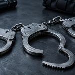 A pair of metal handcuffs lies on a dark, textured surface next to a flashlight and a piece of tactical equipment.