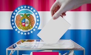 A voter drops a ballot into a ballot box, with the Missouri state flag in the background.