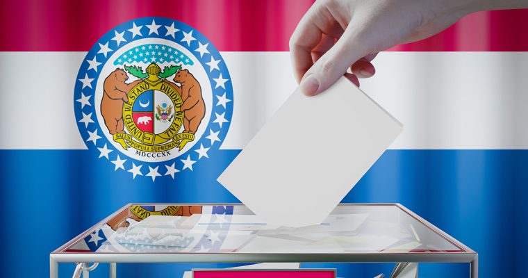 A voter drops a ballot into a ballot box, with the Missouri state flag in the background.