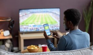Back view of man holding smartphone with sports bets app on screen while watching football match at home
